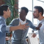 Office conflict. Angry multiracial young men fighting at workplace, afro guy standing between them